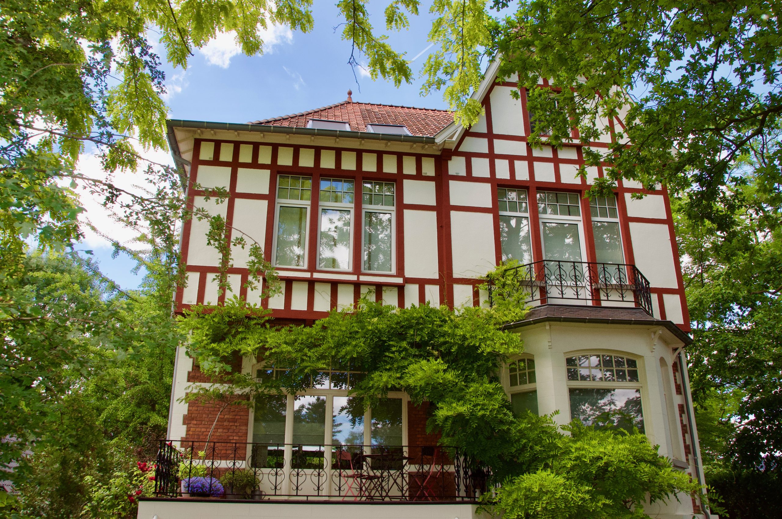 exterior image of the building of Attic 126, The building is white with dark red accents around the windows and roof. On the walls several branches of green nature are sprouting.